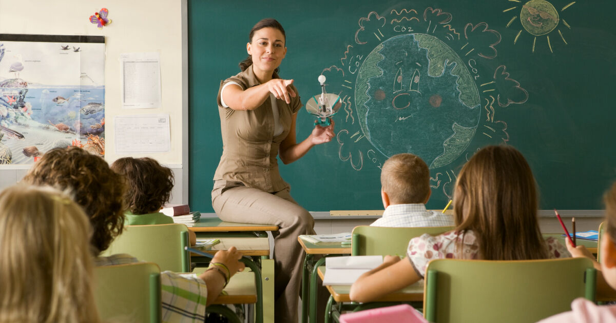 A teacher sits on top of a desk in front of her class. She points at a student.